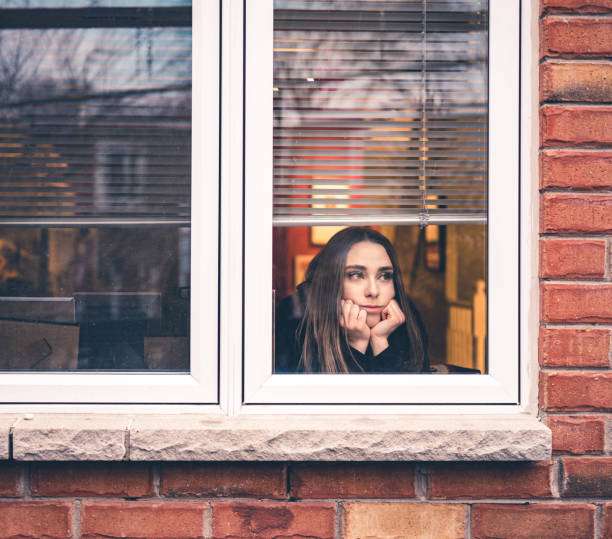 COVID-19, stay home Young woman sitting inside her home, close to the window looking out. She is practicing social distancing due to pandemic of COVID-19 and stay at home order. stay at home order stock pictures, royalty-free photos & images