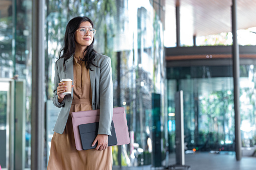Happy Successful Asian Businesswoman Holding a Takeaway Coffee Cup and Files on the Street Next to a Glass Building