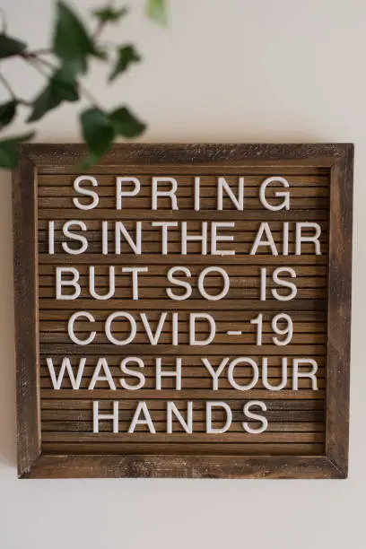 A letterboard with the words "Spring is in the Air, but so is Covid-19. Wash Your Hands." A fun and happy way of saying the main thing we need to do during the Coronavirus. A lush green English Ivy plant in the photo as well.