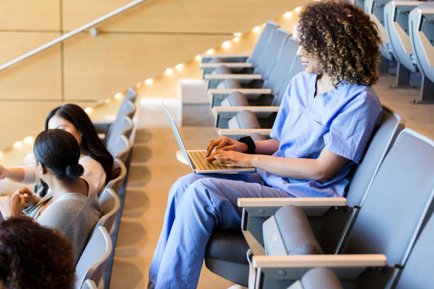 Female student sitting alone in classroom takes notes on laptop The female student, wearing her scrubs, sits alone in a row in the lecture hall and takes notes on her laptop. community college stock pictures, royalty-free photos & images