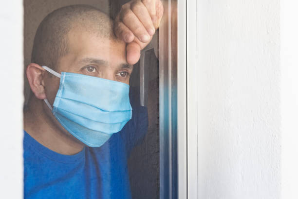 Sick man with face mask looking out the window being quarantined at home stock photo
