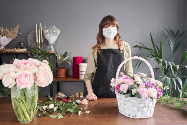 mujer florista con una máscara médica para protegerse del coronavirus. concepto pandémico, trabajando durante una epidemia. pequeña floristería. entrega de flores. - florist women bouquet spring fotografías e imágenes de stock