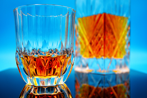 A glass of whiskey straight-up with a decanter on a reflective surface against a blue background in studio.
