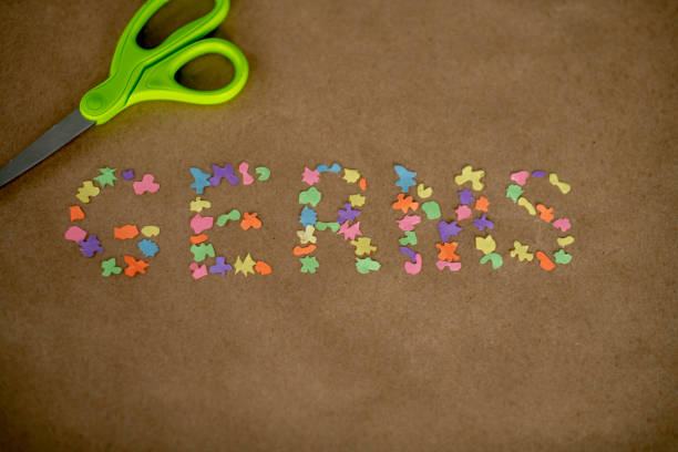 The word GERMS is spelled out in brightly colored pieces of paper, that are cut out in different shapes.  They are laid out on a brown paper background and a pair f green scissors can be seen above.