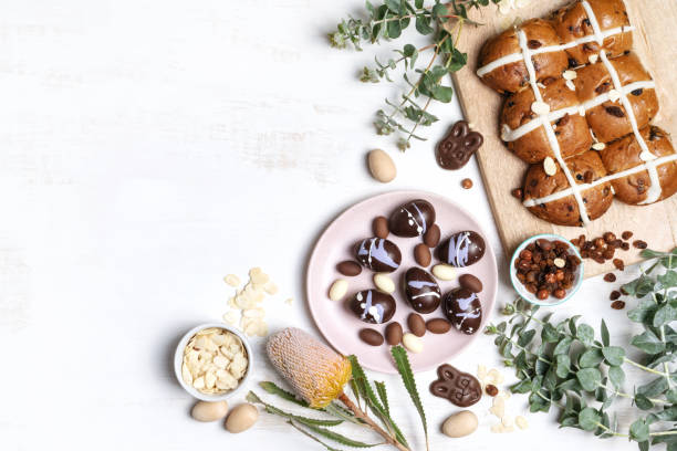 Vegan chocolate Easter eggs and hot cross buns. Vegan chocolate Easter bunny, Easter eggs and homemade hot cross buns on a wooden serve board. Decorated by shaved almonds, hazelnuts, wooden eggs, Australian Banksia and eucalyptus leaves on a white background. sweet bun stock pictures, royalty-free photos & images