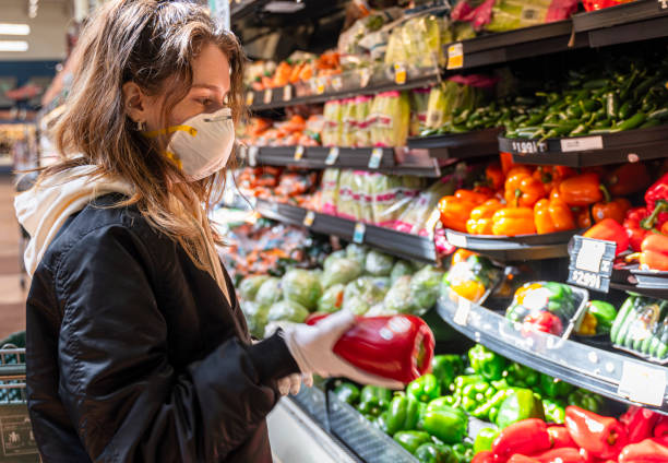 shopping durante una pandemia. una giovane donna che indossa una maschera protettiva e guanti che fanno la spesa di verdure in un negozio. - mature women isolated mature adult women foto e immagini stock