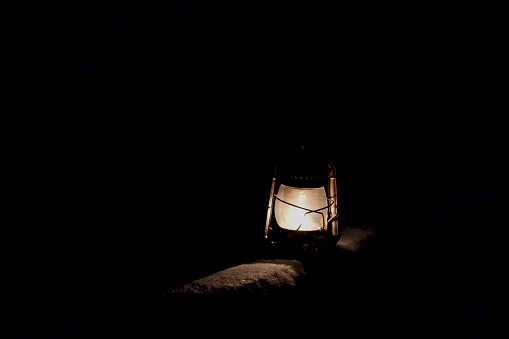 This photo contains an older style kerosene lamp, lit in the dark. It's light glistens off of the surrounding snow.