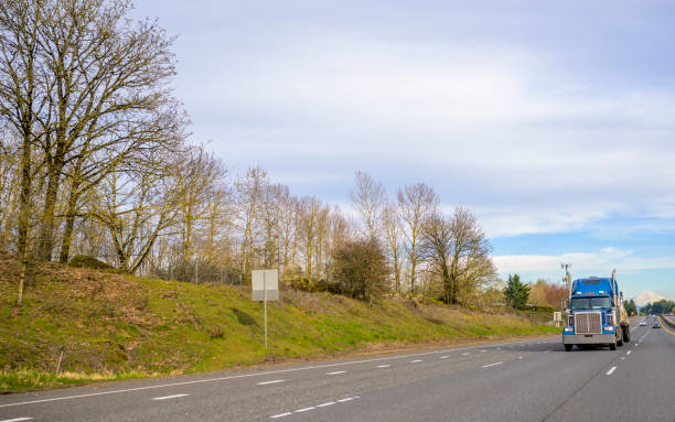 istock Classic powerful blue big rig semi truck transporting covered cargo on flat bed semi trailer running on the wide highway road with trees on the hillside 1215266573