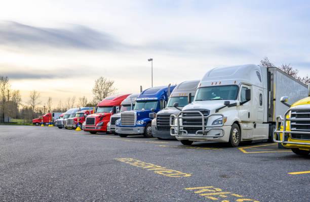 Different big rigs semi trucks with semi trailers standing in row on truck stop parking lot with reserved spots for truck driver rest and compliance with established truck driving regulations stock photo