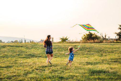 Young Caucasian mother and her daughter running on a meadow with kite.