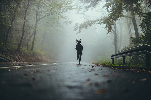 Back view of determined athletic woman jogging during bad weather conditions in forest. Copy space.