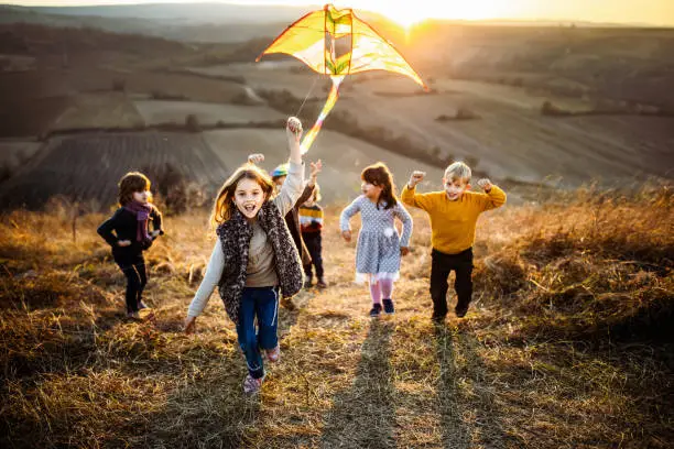 Large group of carefree kids running with a kite up the hill in autumn day. Focus is on girl with a kite looking at camera.