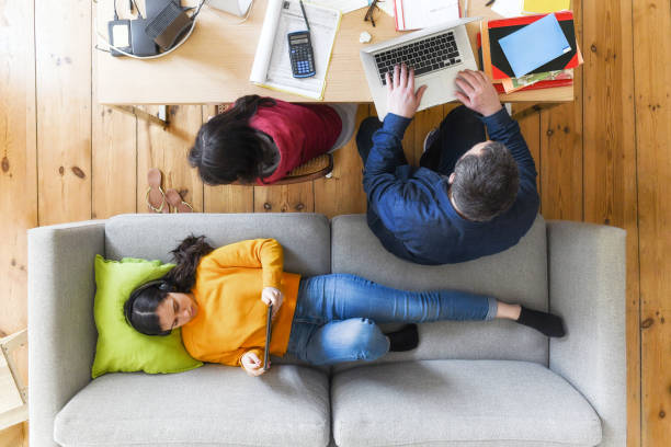 time for home office and home schooling overhead view on man working at table in living room beside daughter doing home work, second daughter looking at tablet on sofa lockdown business stock pictures, royalty-free photos & images