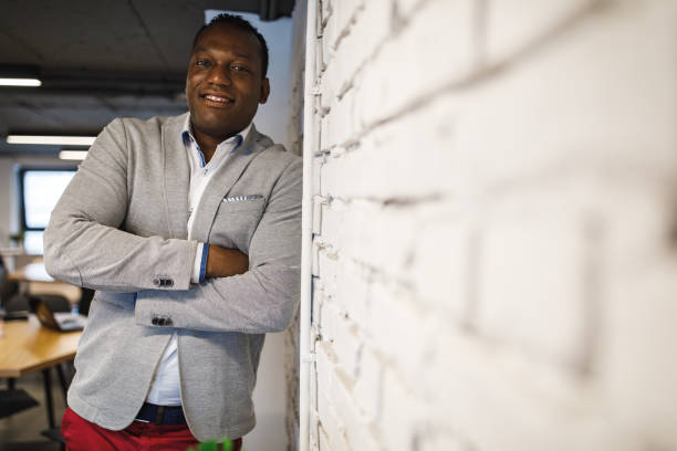 Selective focus of adult Afrolatino businessman in smart casual clothes leaning on a wall at his workplace and inspiring confidence and trust with his smile.