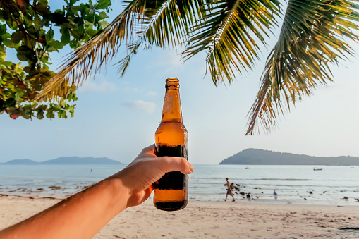 Sunny beach and beer bottle in hand of happy tourist during holidays. Ocean view with fresh beer of a vacationist under palm trees.