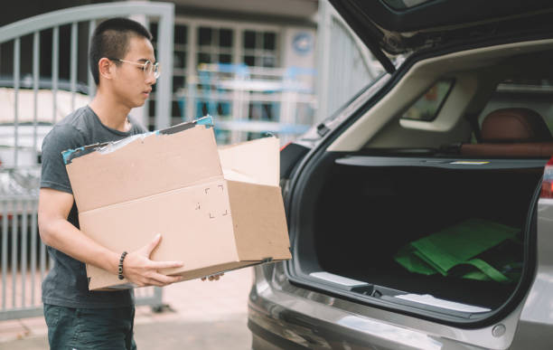 un ragazzo adolescente cinese asiatico che porta un cartone scatola di carta al bagagliaio dell'auto suv di fronte a casa sua - car rear view behind car trunk foto e immagini stock
