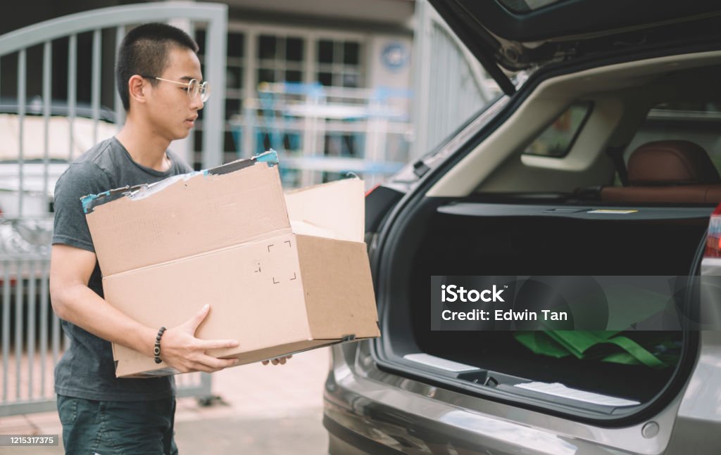 an asian chinese teenager boy carrying a paper box carton to the suv car trunk in front of his house Box - Container Stock Photo