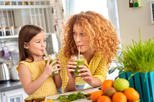 Young mother and her little child girl standing on kitchen together, enjoying healthy smoothie beverage, drink and enjoy fresh coctail