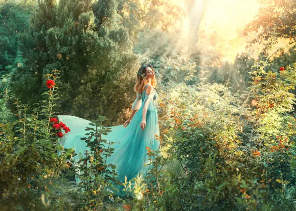 Photo of Fairytale nymph enjoy bright sun in green forest. Hairstyle decorated blue flowers flowing hair. Fairy in long blue airy dress train fly wavy. Backdrop natural tree bushes of red roses divine sunbeams