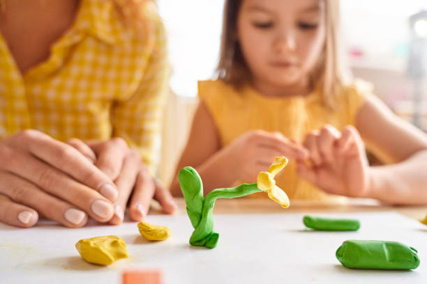 Young mother and her junior daughter playing with plasticine Selective focus on plasticine flower against blurred background with young mother and her junior daughter spending day together, playing at home childs play clay stock pictures, royalty-free photos & images