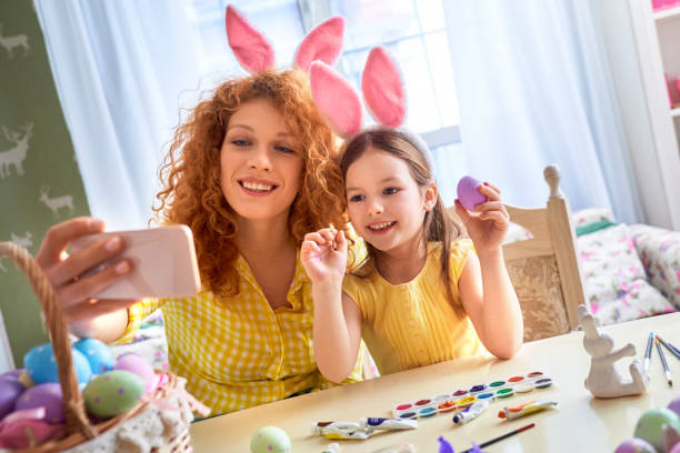 Easter Concept. Mother and daughter in rabbit ears sitting at kitchen coloring eggs taking selfie on smartphone smiling cheerful