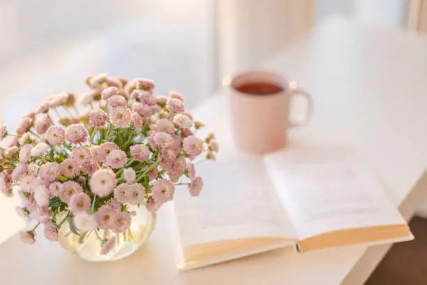 Photo of small pink flowers bouquet in glass vase with blurred soft focused book and pink cup of tea