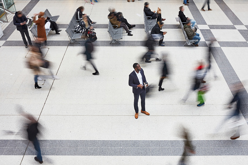 High angle view of 40 year old African businessman standing still in London transportation building while travelers walk by him in blurred motion.