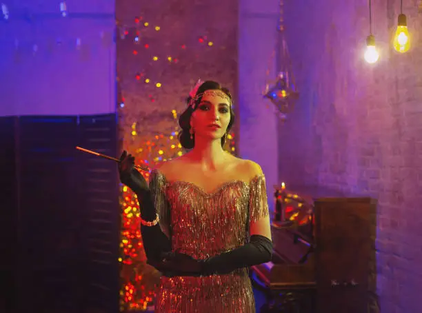 Sexy retro flapper woman in gold dress. Mouthpiece cigarette in hand. Creative neon light roaring party Great Gatsby style. Backdrop classic room bokeh sparkles smoke. Hairstyle finger wave headband