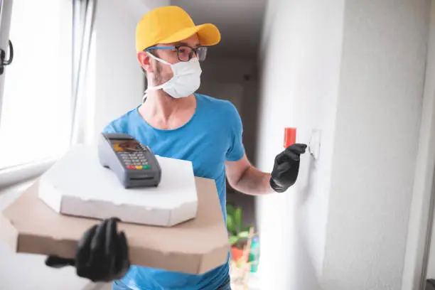 Photo of Deliveryman with protective medical mask holding pizza box and POS wireless terminal for card paying, ringing at the doorbell - days of viruses and pandemic, food delivery to your home and safety hygiene measures.