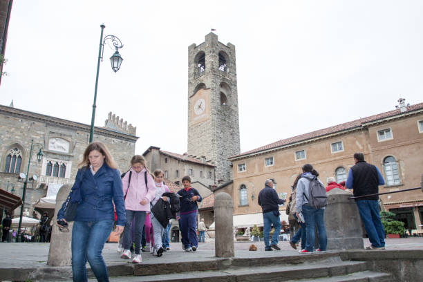 People on and leaving square Piazza Vecchia in Bergamo seen from street some steps below. A woman and some children are walking down steps. In background are tower Campanone and palace Palazzo della Ragione.