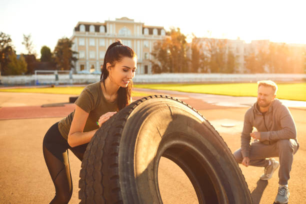 Athletic girl pushes a tire in the stadium at sunset. Man and woman doing gym outdoors. Athletic girl pushes a tire in the stadium at sunset. Sports people. crossfit girl stock pictures, royalty-free photos & images