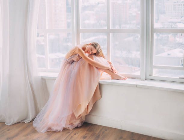 Artwork sleeping dream beauty. Blonde young woman with long flowing hair. sits on windowsill hugs legs. vintage room white window. Princess cute face Girl. luxury pastel beige purple graduation dress Artwork sleeping dream beauty. Blonde young woman with long flowing hair. sits on windowsill hugs legs. vintage room white window. Princess cute face Girl. luxury pastel beige purple graduation dress pink gown stock pictures, royalty-free photos & images