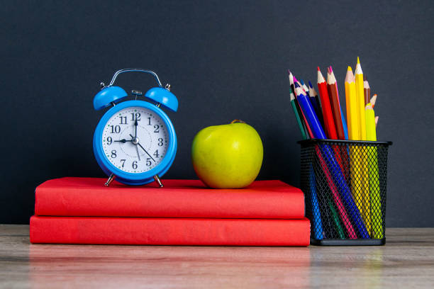 istock The blue alarm clock along with multi-colored pencils stand on a stack of red books, next to an apple 1215342151