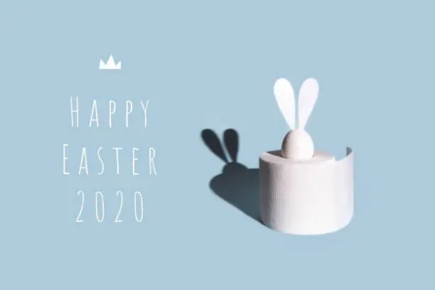 White egg with hare ears in a roll of toilet paper. Hard shadow on a light background. Concept on Easter 2020