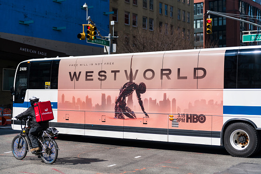 New York City, USA - March 19, 2020: During a state of emergency is New York City due to the growing coronavirus pandemic, a food delivery person for Chowbus rides his bike adjacent to a New York City bus advertising the latest season of HBO's science fiction series Westworld.