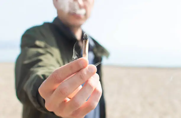 Photo of Young man holding a lit marijuana joint while smoking on the beach. Blur background and copy space right.