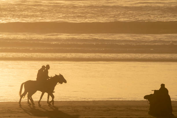Camel, horses and Berber tribesman at Taghazout, Morocco. With the sun reflected on the sea and silhouetting them against the waves. There are tourists on the horses and a tribesman with a camel in the golden unfiltered rays of the setting sun.