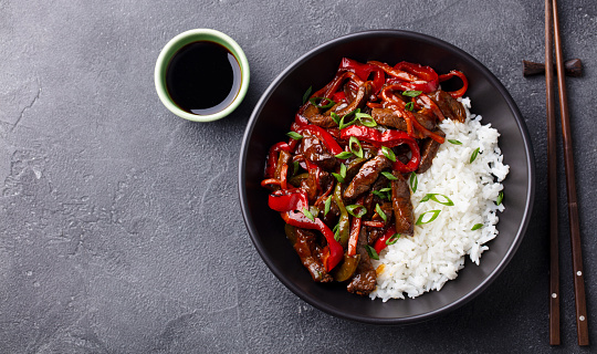 Beef and vegetables stir fry with white rice in a black bowl. Grey background. Copy space. Top view.