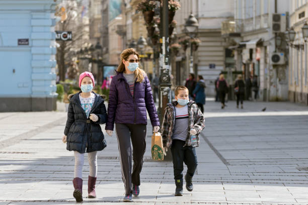 Mother and two young kids walking on the city streets wearing face masks against coronavirus. COVID - 19 pandemic. Street view in city center stock photo