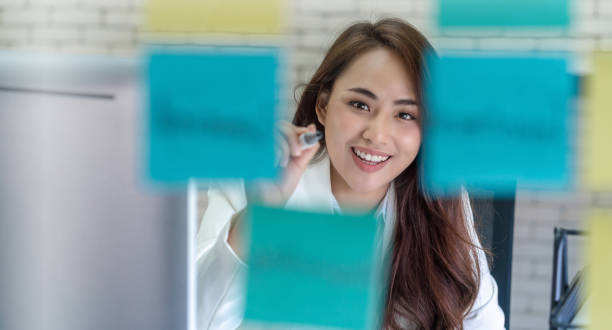Asian Businesswoman writing the post it on the mirror Asian Business woman concentrating and writing the post it on the mirror in happiness action at office mirror writing stock pictures, royalty-free photos & images