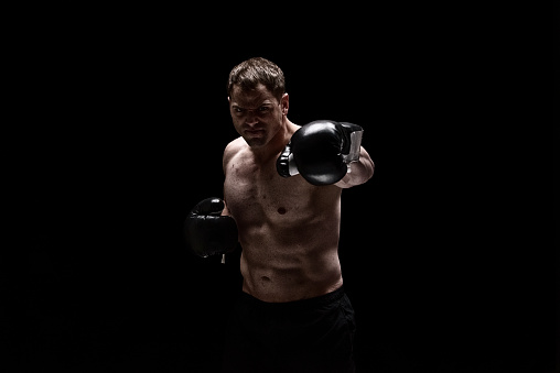 Front view of with brown hair caucasian male in front of black background wearing glove who is serious and boxing and showing fist who is punching