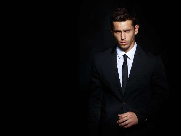 Handsome young brunette model, wearing in black and white suit, looking with attitude, posing at studio, isolated on black background. Business man portrait. Copy space. Horizontal view. Image of a confident brunette man in black suit, isolated in a black background. hot handsome man stock pictures, royalty-free photos & images