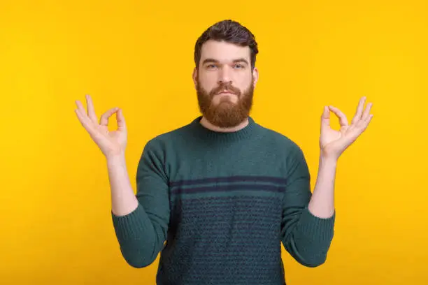 Serious bearded man is trying to practice yoga  and reach zenon yellow background.