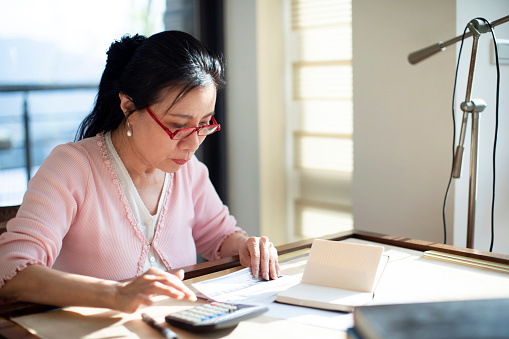 Senior Asian woman sitting at desk at home and working with calculator and documents
