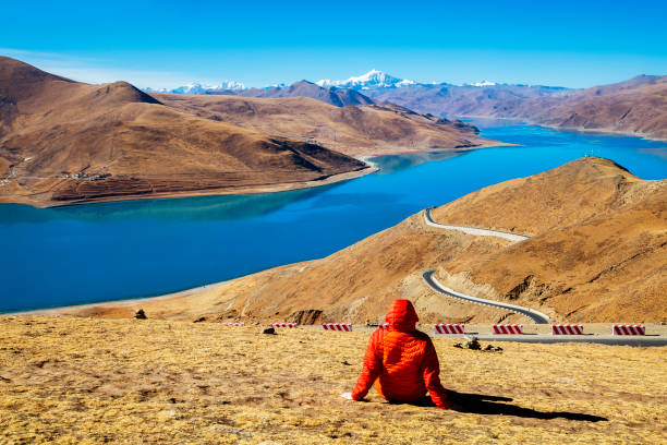 Rear view of female hiker sitting at a height of 4900 m in Tibet. In the background is seen Yamdrok Lake (Yamdrok Yutso in Tibetan dialect) and the Noijinkangsang peak (7206 m) in Himalaya. Yamdrok Lake is situated about 100 km from Lhasa, the capital of Tibet.