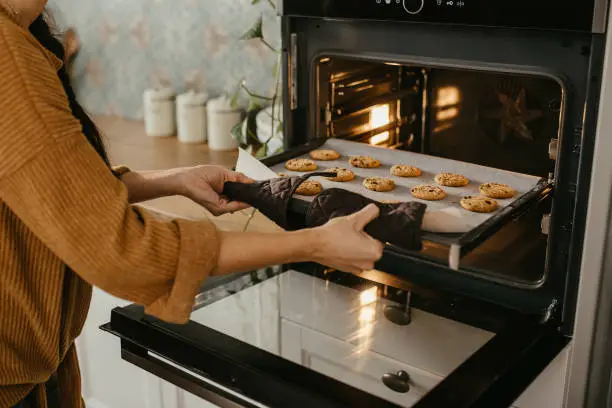 Young mother is putting a tray full of cookies which they made together with her husband and her son in the oven. Horizontal photo. Face is not visible.