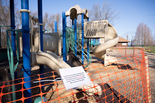 Playground closed to prevent spread of COVID-19. An orange fence is placed around a children's playground at a public park. A sign reads \
