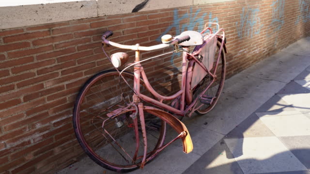 Cute vintage pink bicycle at the street leaning on the red brick wall with painted blue graffiti. Abandoned retro bicycle with broken wheel outdoors