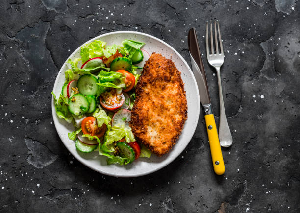 chicken milanese and fresh romaine salad, cherry tomatoes, radishes, cucumbers salad - delicious lunch on a dark background, top view - milanese imagens e fotografias de stock