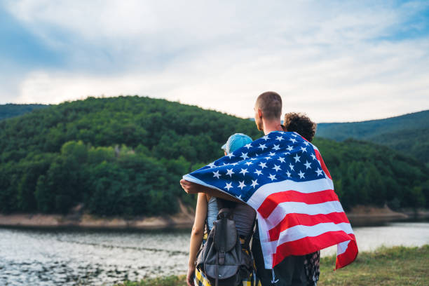 Small caucasian group of young people walking in nature near lake and forest and proudly showing American flag.